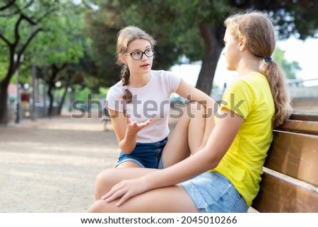Youth girls in shorts sitting on bench in city park and discussing something.