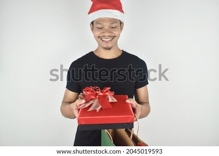 Portrait of young man in christmas hat smiling holding red gift box and shopping bag. Christmas and New Year holiday concept. 
