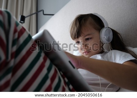 Little girl plays in the tablet while lying on the bed. The child listens to music on headphones. Incorrect position of the spine while playing with the gadget. Violation of the foundation.