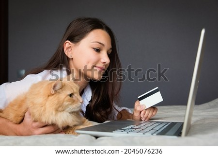 a girl with a red cat is lying on the bed with a laptop and holding a credit card in her hand