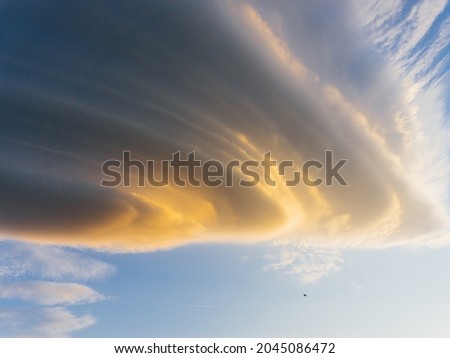 Gray and orange swirling clouds in a clear blue sky in the shape of a funnel mixed with the rays of the setting sun.