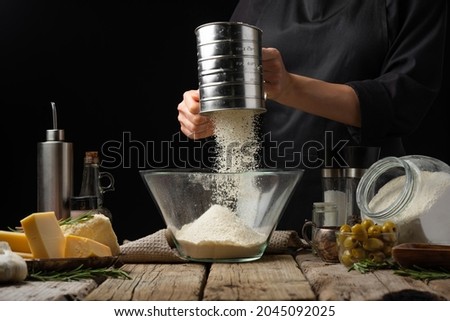 The chef sifts the flour for the dough through a sieve into a large glass bowl. Ingredients for making focaccia, pizza, pie. Black background, wood texture. Restaurant, hotel, cafe.