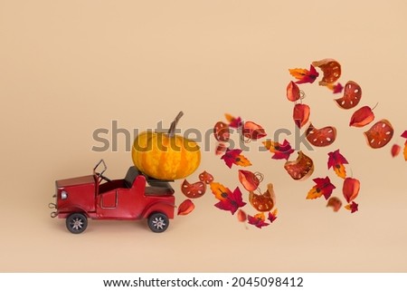 Red old car carrying big pumpkin and swirling leaves against minimalistic beige background. Creative minimal Halloween or Thanksgiving season concept. Copy space