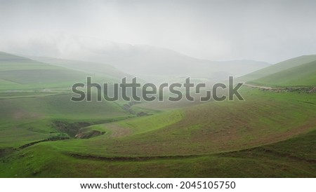 The copter's flight in the afternoon over a green gorge towards the hills and a cloudy rainy sky. A beautiful idyllic landscape with green fields, roads and hills on a cloudy day. Beautiful natural. Royalty-Free Stock Photo #2045105750