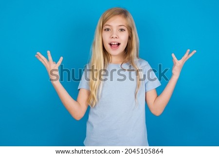 Joyful excited lucky beautiful caucasian little girl wearing blue T-shirt over blue background cheering, celebrating success, screaming yes with clenched fists