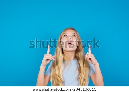 beautiful caucasian little girl wearing blue T-shirt over blue background being amazed and surprised looking and pointing up with fingers showing something strange.