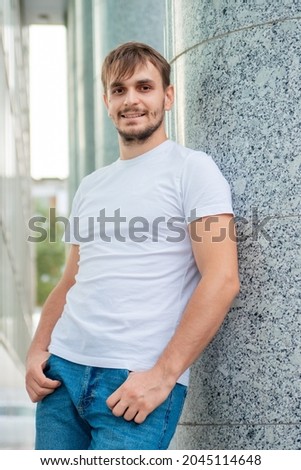 A young guy in a blank white T-shirt stands against the background of a gray column. A man near an office building in the city. Mock-up.