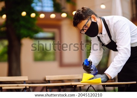 Disinfecting to prevent COVID-19. Waiter cleaning the table with Disinfectant Spray in a restaurant wearing protective medical mask and gloves.