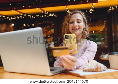 Young woman in casual clothes sit at table in cafe shop eat breakfast using laptop pc computer take photo of credit bank card by mobile phone shopping online order delivery relax in restaurant indoors