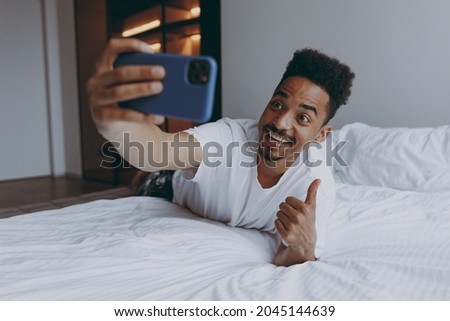 Full length fun young african american man in nightwear do selfie shot on mobile cell phone show thumb up gesture lying in bed rest relax spend time in bedroom lounge home in own room house wake up