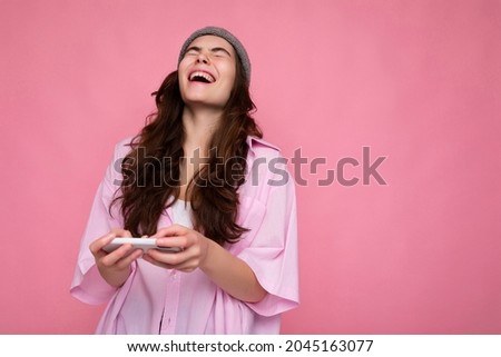 photo of attractive positive laughing young brunette woman wearing stylish pink shirt and grey hat isolated over pink background holding in hand and using mobile phone playing online games and having
