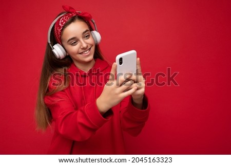 Positive smiling attractive brunette girl wearing red hoodie isolated on red background holding and using smartphone reading online wearing white wireless headphones listening to music looking at