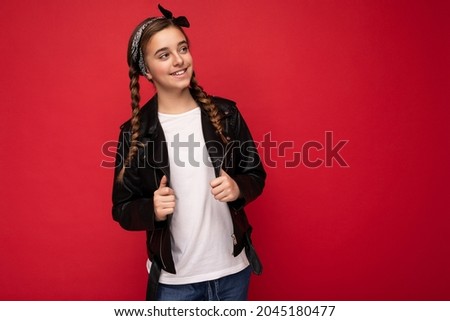 Photo shot of beautiful happy smiling brunette little girl with pigtails wearing trendy black leather jacket and white t-shirt for mockup standing isolated over red background wall looking to the side