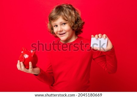 Portrait photo of positive happy smiling little boy with curly blond hair with sincere emotions wearing casual red pullover isolated over red background with copy space, holds red piggy box for money