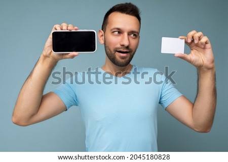 Photo of handsome young unshaven brunet man with beard wearing everyday blue t-shirt isolated over blue background holding and using mobile phone and credit card making payment looking to the side