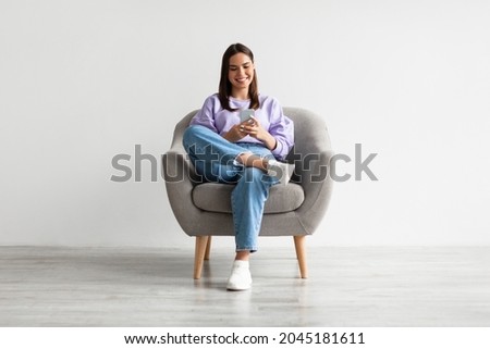 Beautiful young woman sitting in armchair, looking at mobile phone screen, using new cool application against white studio wall, full length. Joyful lady using smartphone for work or entertainment