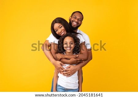 Healthy Family Relationship. Portrait of cheerful African American man hugging his wife and daughter from behind back standing isolated on yellow orange studio wall, posing looking at camera
