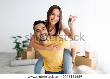 Cheerful Caucasian lady getting piggyback ride from her Arab boyfriend, showing house key indoors. Lovely multinational couple posing in new apartment on moving day Royalty-Free Stock Photo #2045181659
