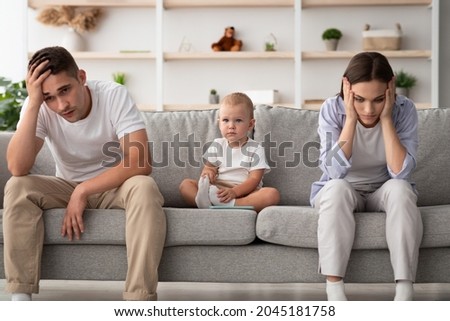 Family Conflicts. Cute infant baby sitting between offended parents after quarrel at home, young spouses with child suffering marital crisis, feeling depressed and frustrated, free space Royalty-Free Stock Photo #2045181758