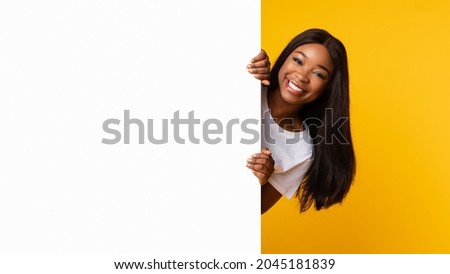 Intresting Offer. Happy Casual Black Woman Peeping Out The Side Of White Advertisement Board For Text Or Design. Smiling Cheerful Lady Holding Billboard, Looking At Camera, Standing Over Yellow Wall