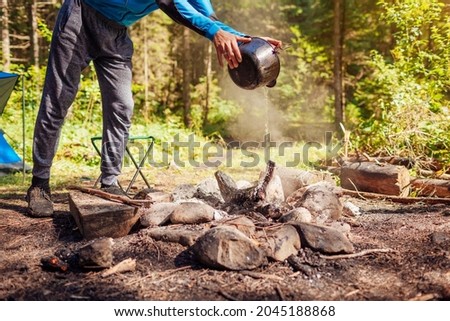 Man extinguishing campfire with water from cauldron in summer forest. Put out campfire by tent. Traveling fire safety rules Royalty-Free Stock Photo #2045188868
