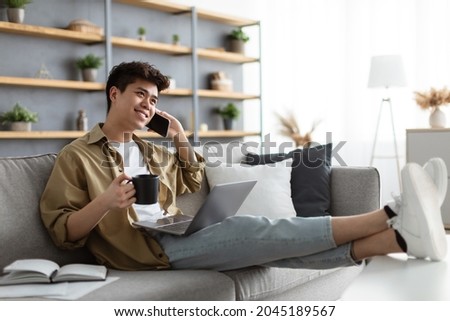 Cheerful Asian Freelancer Guy Drinking Coffee And Talking On Cellphone While Having Break During Work With Laptop Computer At Home Office, Speaking With Friends, Resting Sitting On Sofa, Free Space Royalty-Free Stock Photo #2045189567