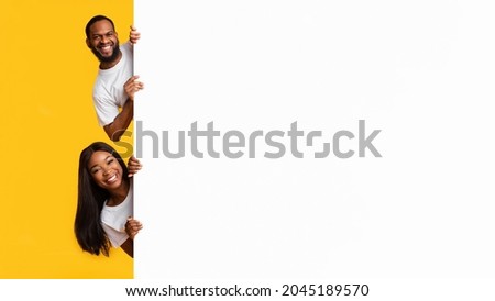 Intresting Offer. Happy Casual Black Couple Peeping Out The Side Of White Advertisement Board For Text Or Design. Smiling Cheerful People Holding Billboard, Looking At Camera, Standing On Yellow Wall Royalty-Free Stock Photo #2045189570