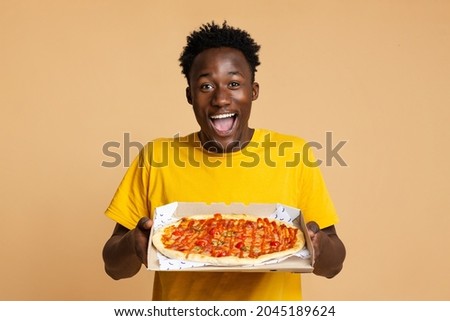 Happy Excited Black Guy Holding Opened Box With Tasty Italian Pizza While Standing Isolated Over Beige Studio Background, Cheerful Young African American Man Enjoying Food Delivery Service