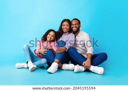Family Day, Parenthood And Childhood Concept. Smiling funny young black parents mom dad sitting on the floor with child teen girl, hugging daughter isolated on blue color background, studio portrait