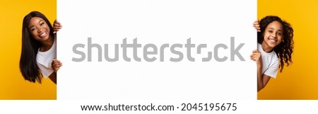 Promotion Billboard Concept. Cheerful African Woman Mom And Girl Holding White Advertisement Board, Standing Together On Yellow Background, Looking At Camera Peeping Out The Two Sides Of Sign Board Royalty-Free Stock Photo #2045195675