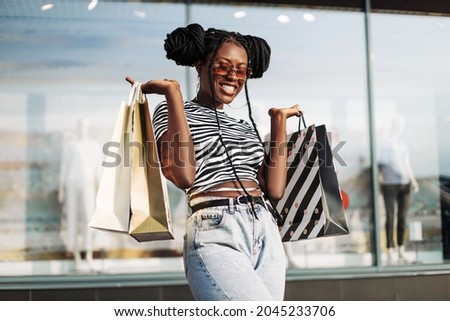 Beautiful curly African American woman, wearing sunglasses, standing in a mall with shopping bags in her hands, enjoying shopping on Black Friday