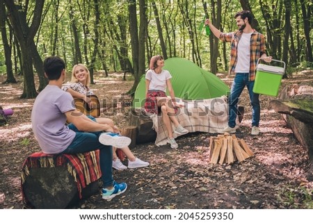 Full length body size photo young friends team staying on camping in summer forest playing guitar drinking beer