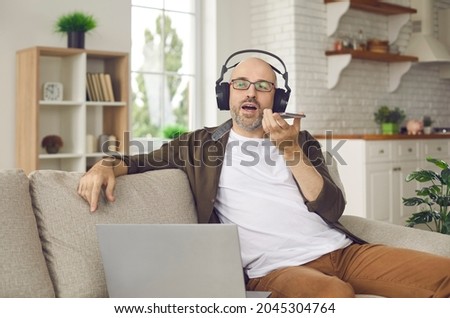 Man sitting on sofa with notebook PC at home, holding mobile, talking on messenger speakerphone. Middle aged bald guy in headphones using audio note taking app or recording work voice message on phone