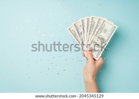 First person top view photo of hand holding fan of money dollars over shiny sequins on isolated pastel blue background with copyspace
