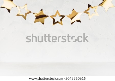 Garland of gold glossy stars hang on string above soft light white wood table, plaster wall. Simple light festival scene for design, presentation, showing.