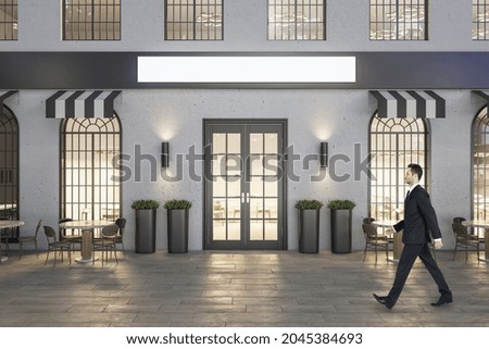Young businessman in suit walking past creative concrete cafe exterior with terrace furniture and empty mock up banner at night