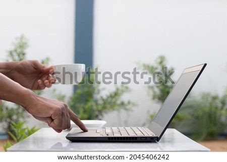 image of working man's hand The left hand is raising a white coffee cup. The right hand is using the index finger to move the keyboard on a black notebook computer. In the backyard, work is relaxing. 