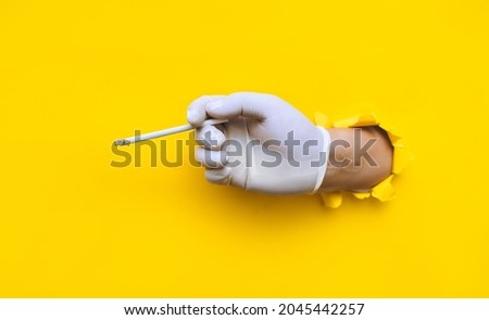 The hand of a doctor (nurse) in a white protective medical glove holds a slims cigarette.Torn hole in yellow paper.The concept of smoking, habit control among health workers during the covid-19 period