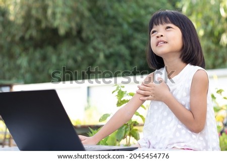 A cute-looking Asian kindergarten girl  is studying online on a black laptop in her backyard. left hand raised on chest. Look upward in a pose that uses the thoughts and imaginations of the class.