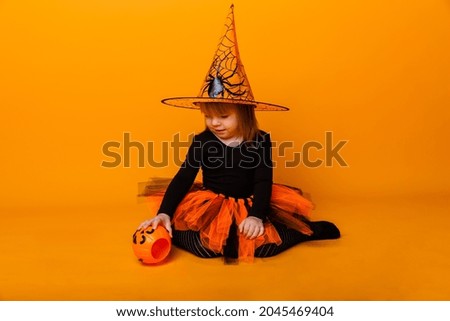 Celebrating Halloween. Little girl dressed as a witch holds a bucket of pumpkins on a yellow background.