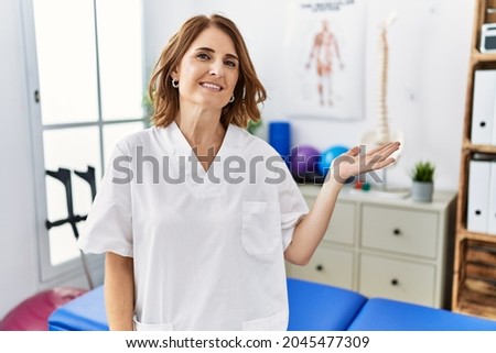 Middle age physiotherapist woman working at pain recovery clinic smiling cheerful presenting and pointing with palm of hand looking at the camera. 