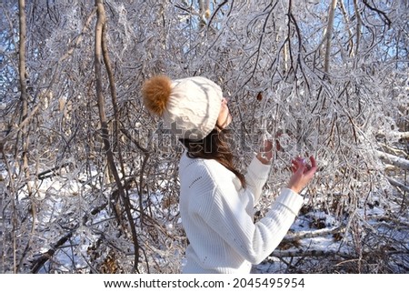 Portrait of young Russian woman in white sweater and beanie next to frozen branches of birch trees, covered with ice. Winter forest in Russia