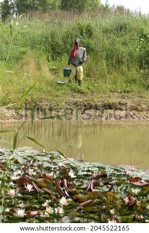 scarecrow dressed in clothes with a gas mask and bucket in his hands, standing on the shore of a pond 