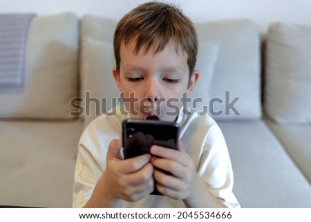 Cropped shot of an excited cute little boy using mobile smartphone while sitting on the sofa at home watching cartoon video or making call playing games entertainment in childhood. Technology concept.