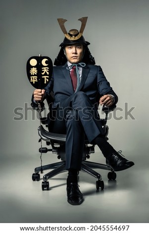 Japanese businessman wearing a samurai's helmet who sits on office chair. Japanese translation: Peace of the world.