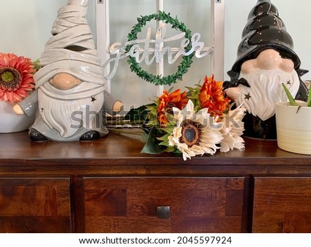 A mantle full of fun Autumn, Thanksgiving, and Halloween decorations. There's a fake windowpane that reads "gather", ceramic gnomes dressed as a mummy and witch, sunflowers, and glass pumpkins.