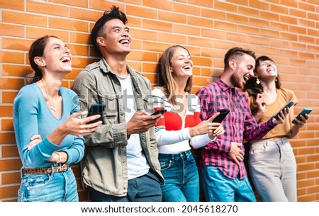 Multicultural friends laughing using smartphone at wall on university college campus - Young people addicted by mobile smart phones - Technology concept with always connected milenials - Vivid filter Royalty-Free Stock Photo #2045618270