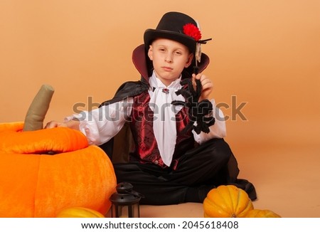 An American boy sits on a beige background next to a huge pumpkin in the costume of Count Dracula or a vampire holding a black big spider on Halloween talking about the holiday waiting for a party