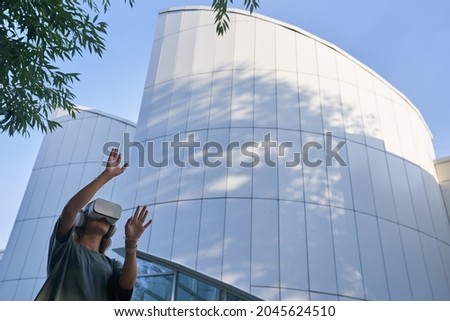 A girl is having fun in virtual reality glasses, standing against the background of a mirrored building. The concept of the future. High-quality photo