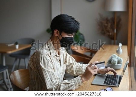 Photo of a young man holding a credit card in hand while using a laptop computer at the wooden counter. Paying by credit card concept.